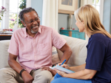 Photo focused on an older man, smiling, talking with a support worker at home.
