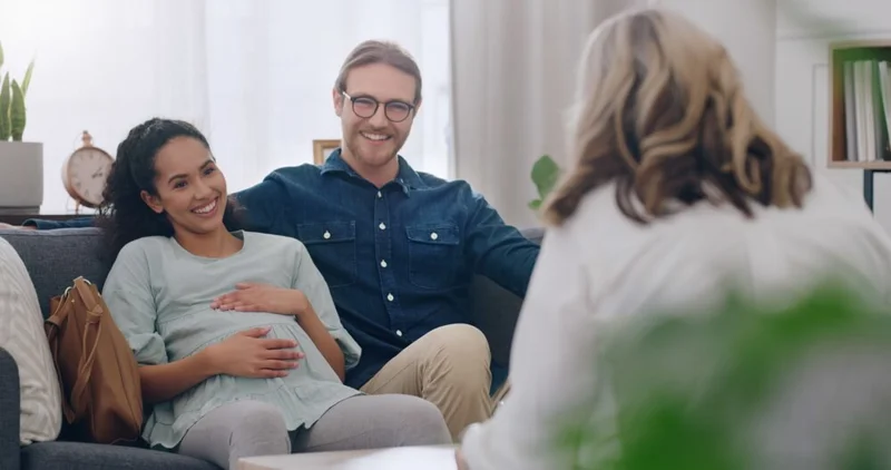 Photo of a pregnant woman, sat next to a man, both smiling, talking to a support worker (whose can be seen from behind in the image).