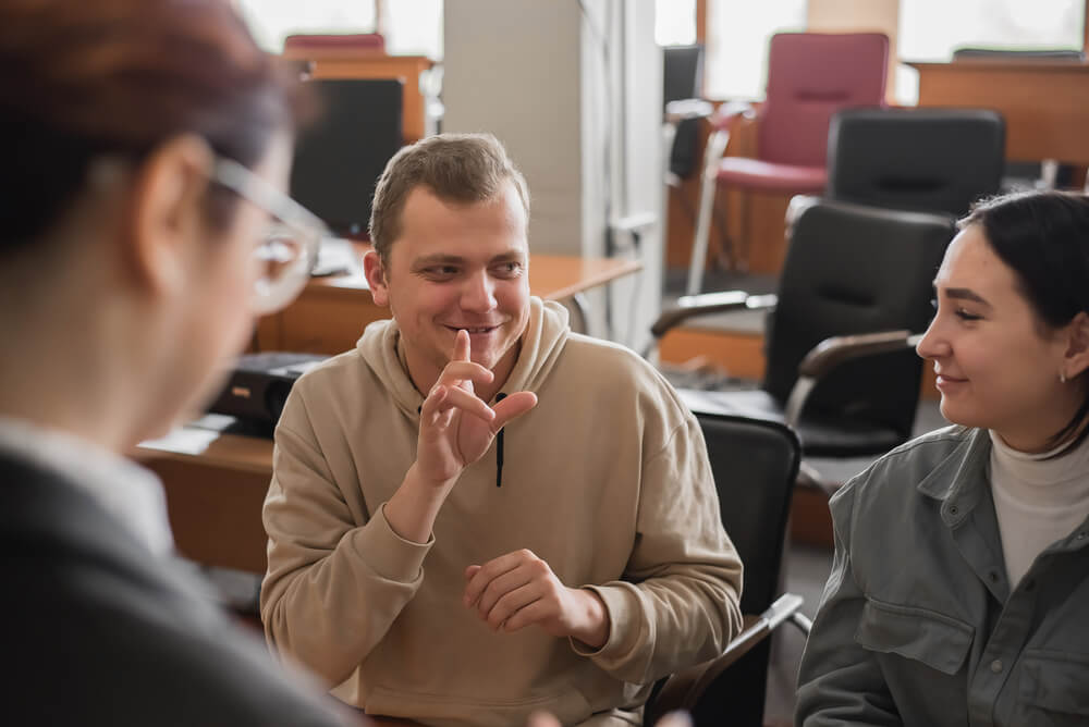 Photo of 3 people signing in a lecture hall. The man in the centre is signing, two women are communicating with him, one blurred out of focus with back to viewer.