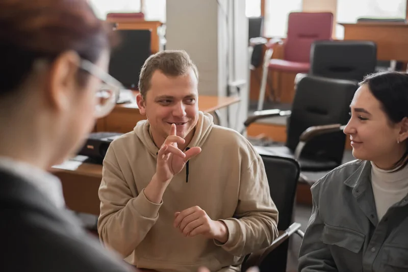 Photo of 3 people signing in a lecture hall. The man in the centre is signing, two women are communicating with him, one blurred out of focus with back to viewer.