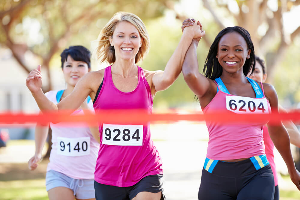 Two women running, crossing the finish line holding hands with their arms raised. Other runners in the background.