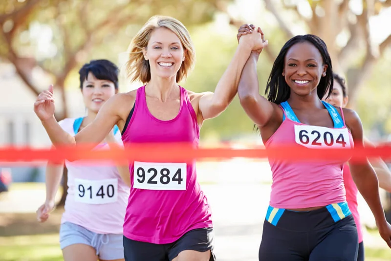 Two women running, crossing the finish line holding hands with their arms raised. Other runners in the background.