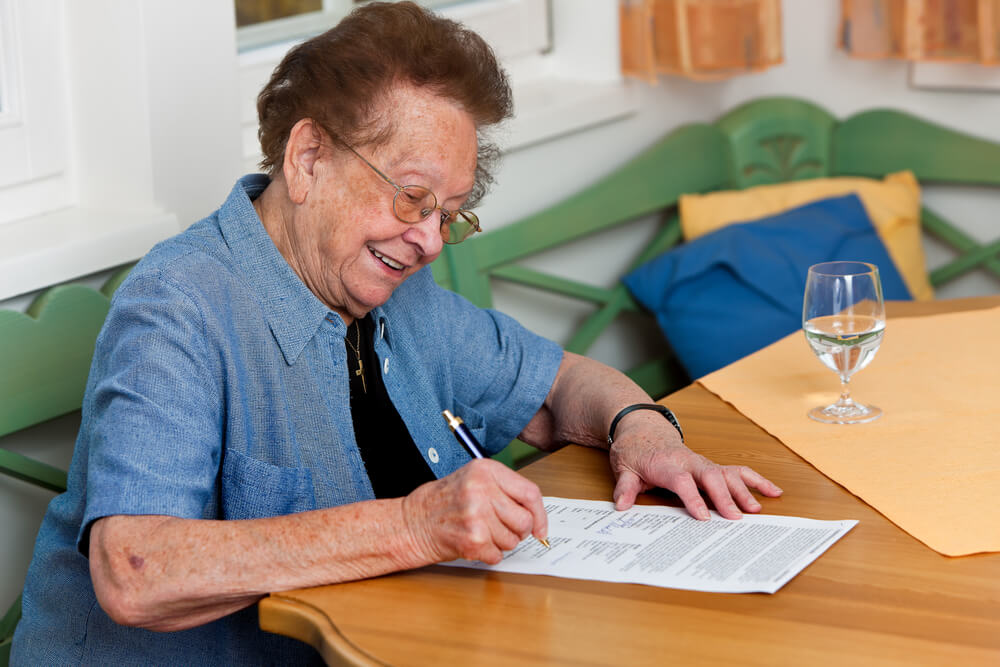 Photo of an older woman sat at a table signing a will.