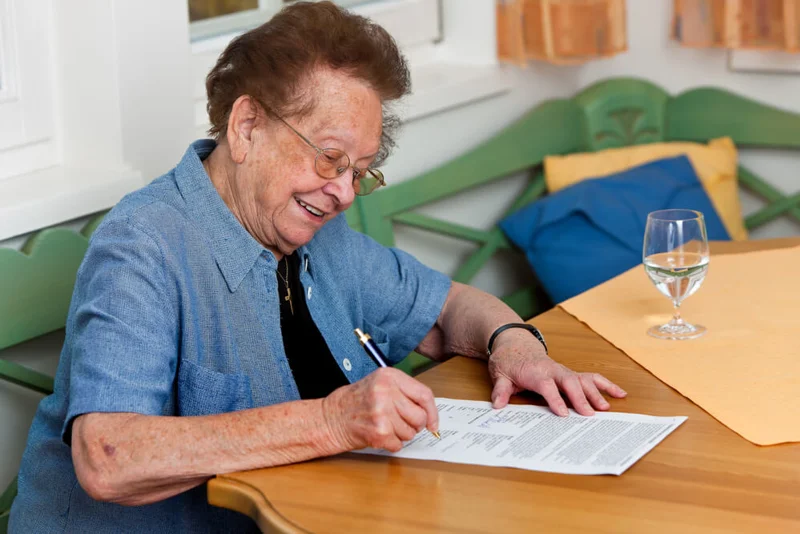Photo of an older woman sat at a table signing a will.