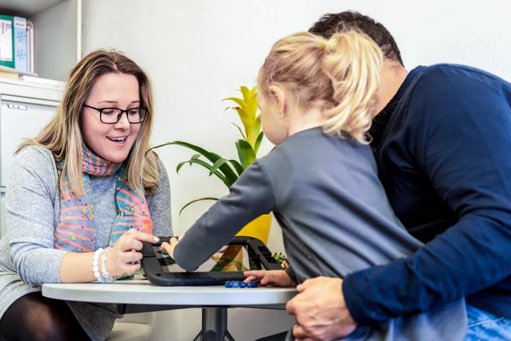 Photo of a support worker, looking at items on a desk with a child and her father.