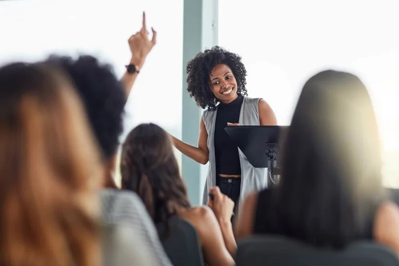 Photo of a group, with raised hands, focused in on a woman at the centre of the image happily answering questions.