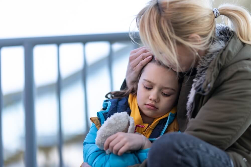 Photo of a mother, sat down in front of railings, with her daughter next to her, resting her head on her mother, who holds her head in her hand. 