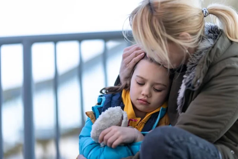Photo of a mother, sat down in front of railings, with her daughter next to her, resting her head on her mother, who holds her head in her hand. 