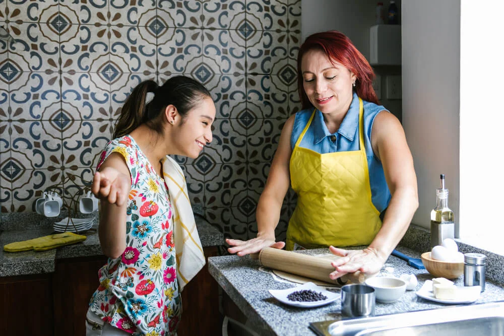 Photo in a kitchen of a young disabled woman baking with another woman, both smiling. 