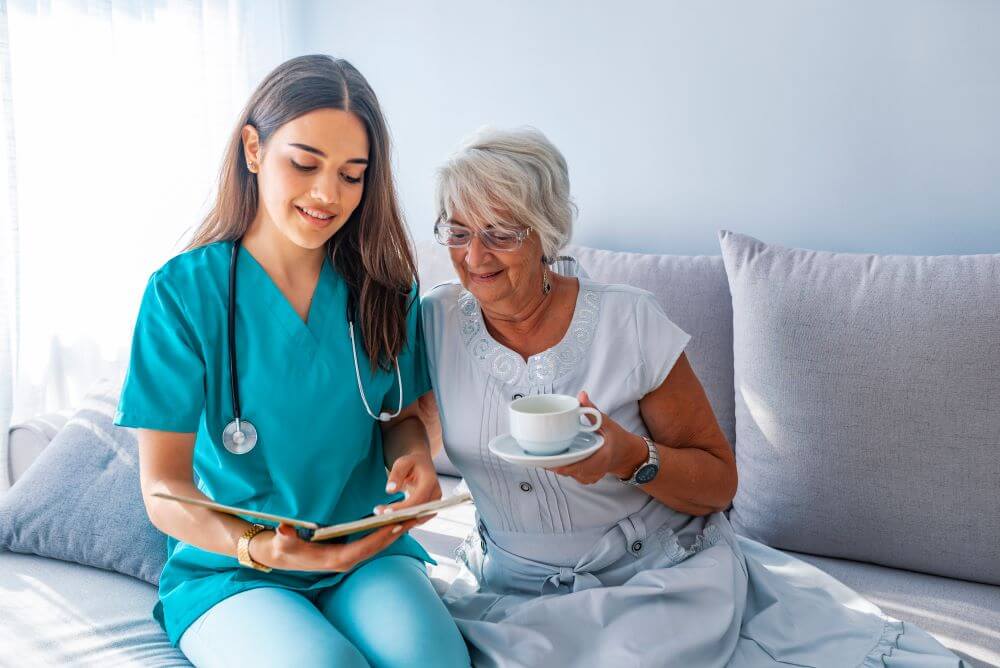 Photo of woman in scrubs sat on sofa with older woman holding cup of tea as they look at a chart together