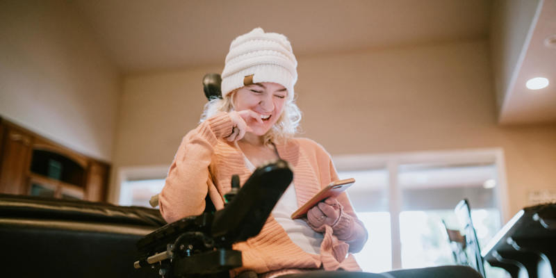 Image of a young disabled woman in a wheelchair, smiling, looking at her phone. Slightly blurred background, indoors.