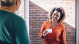 Photo focused on a smiling woman holding up a speakerphone with one hand. In the background there are other people, smiling, but blurred slightly.