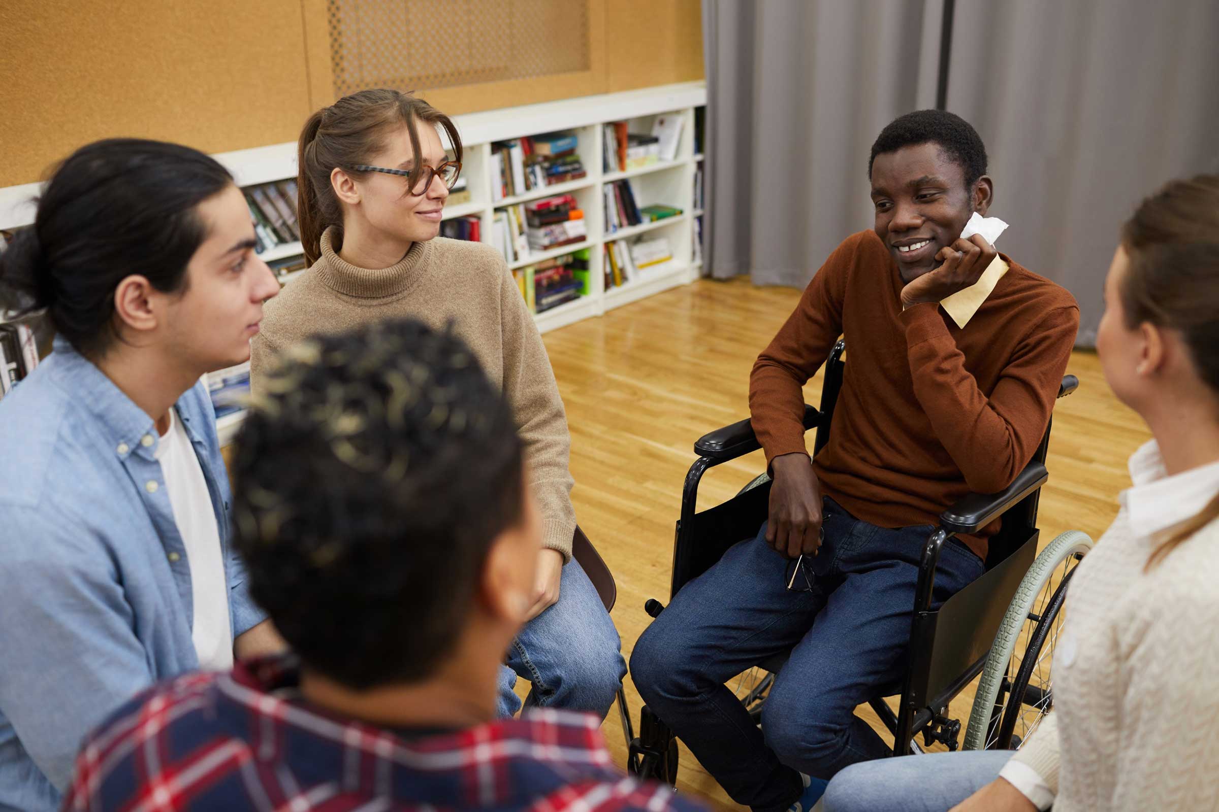 Group of people sat in a circle informally talking, some are blurred. In focus are a man in a wheelchair, a woman, and another man.