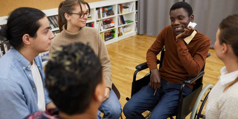 Group of people sat in a circle informally talking, some are blurred. In focus are a man in a wheelchair, a woman, and another man.