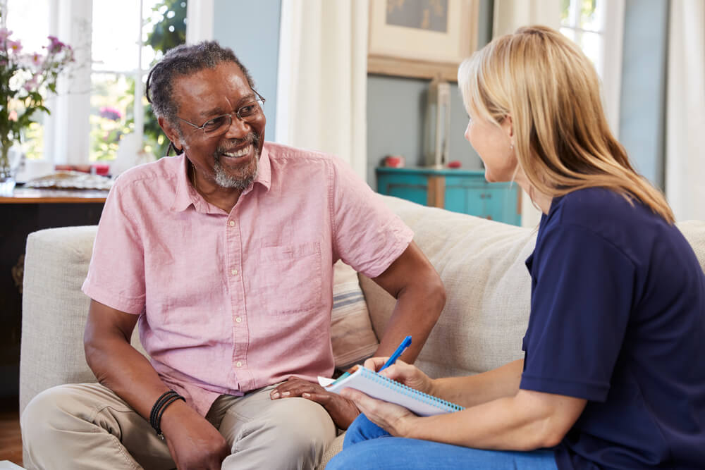 Photo focused on an older man, smiling, talking with a support worker at home.