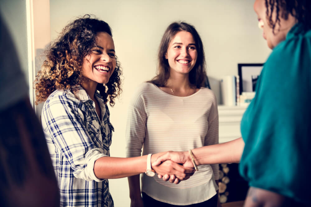 Two women meeting another woman (who has her back to the viewer), shaking hands and smiling.