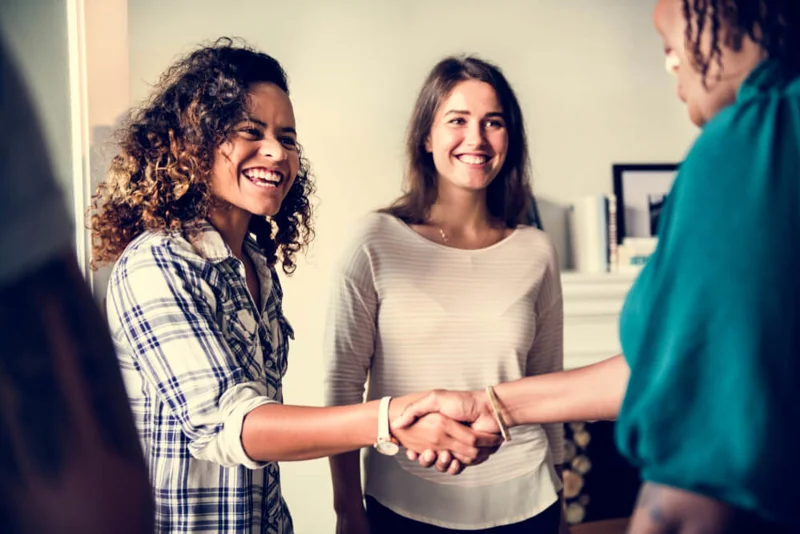 Two women meeting another woman (who has her back to the viewer), shaking hands and smiling.