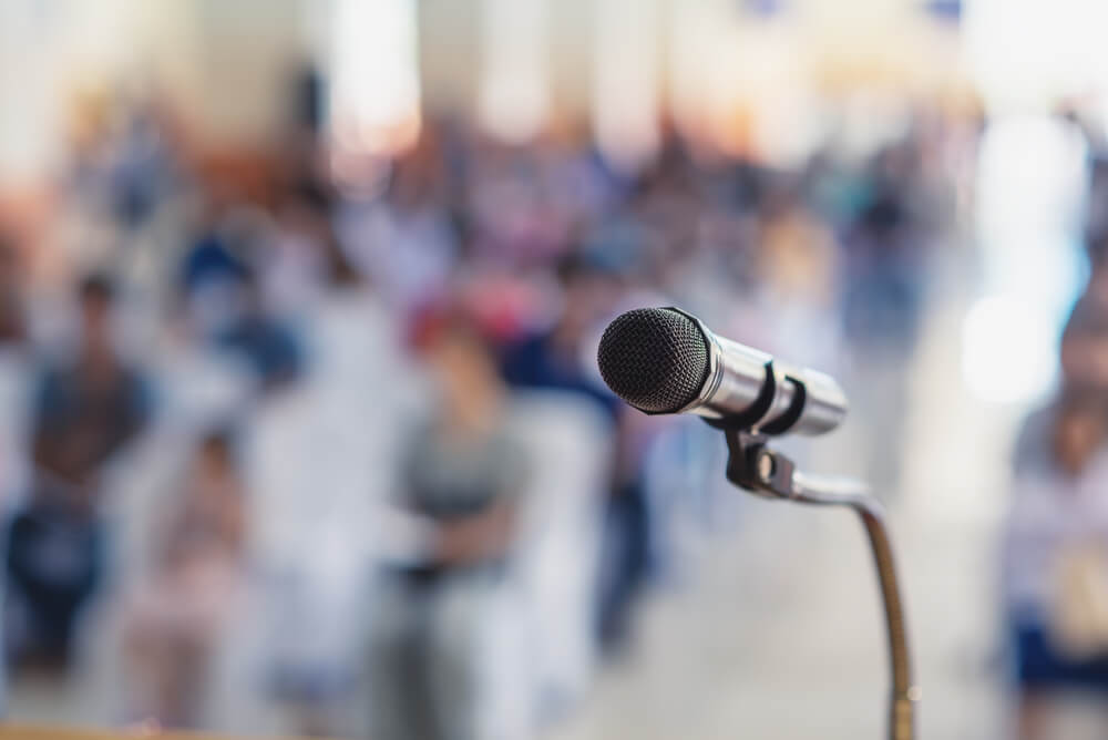 Photo focused in on a microphone, with a blurred background of people listening.
