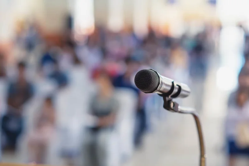 Photo focused in on a microphone, with a blurred background of people listening.