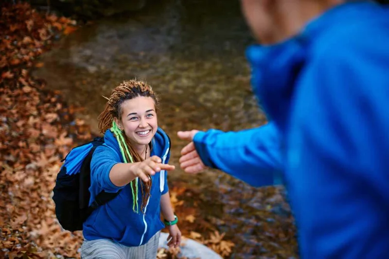 Photo focused on a young woman, smiling, outdoors walking up an incline, hand outstretched to a man reaching for her hand. He is blurred with his back to the viewer.