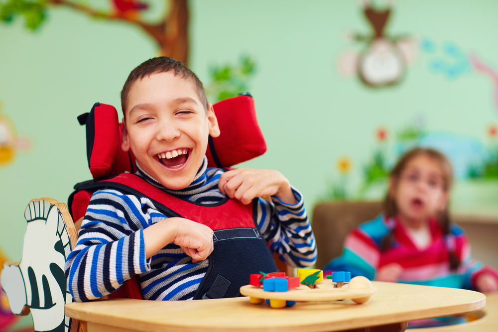 Photo of two disabled children, focused in on a boy, smiling, who is in a wheelchair, and playing with coloured bricks.