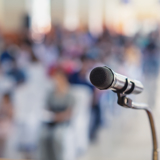 Photo focused in on a microphone, with a blurred background of people listening.