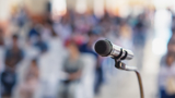 Photo focused in on a microphone, with a blurred background of people listening.