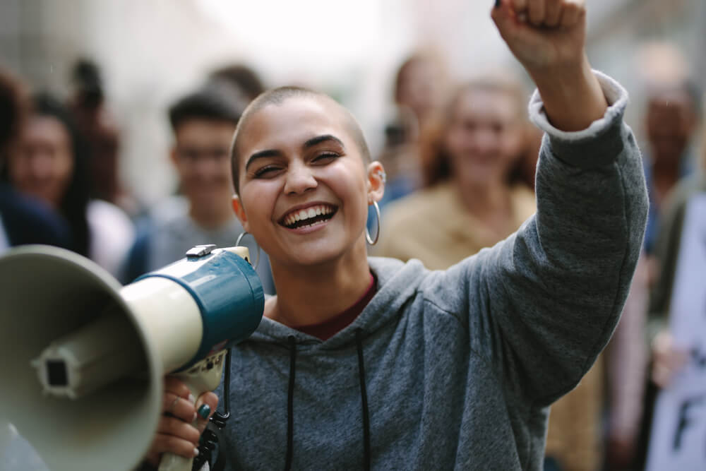 Photo focused on a smiling young woman with a megaphone in one hand and holding her other arm up. The background is blurred and of other people. 