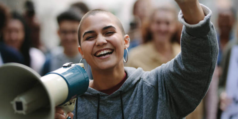 Photo focused on a smiling young woman with a megaphone in one hand and holding her other arm up. The background is blurred and of other people. 