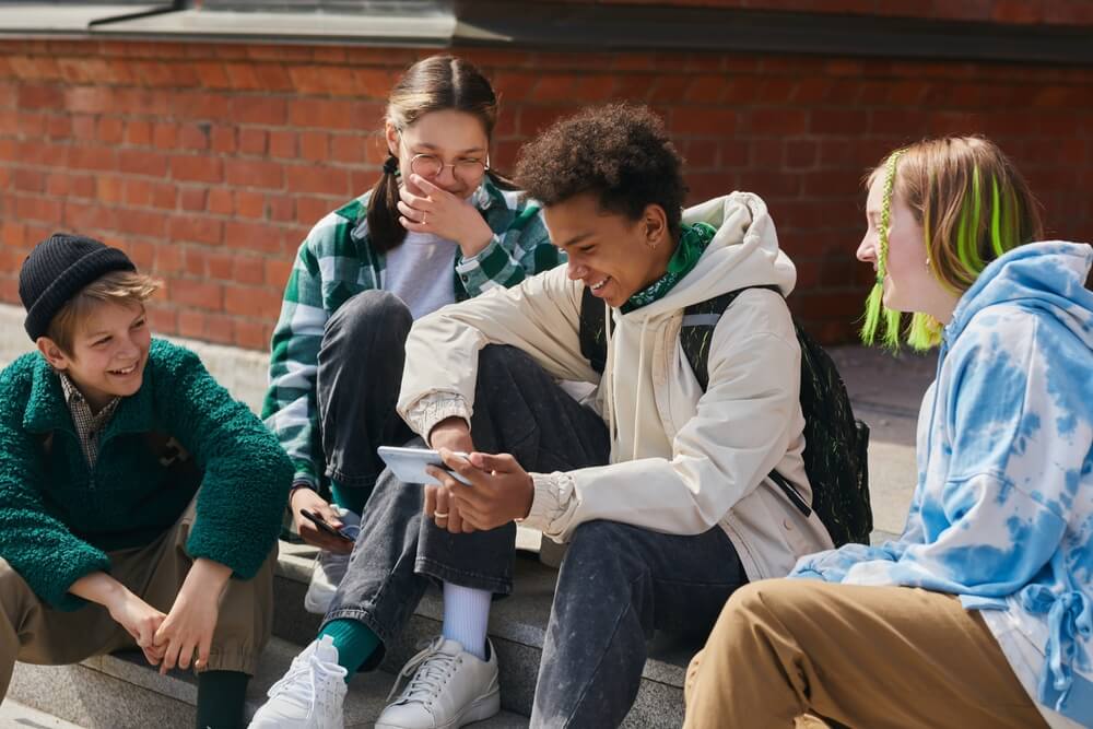 Group of 4 teenagers sitting together outside, chatting, looking at a phone