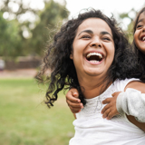 Photo of a mother and daughter outdoors, smiling, the child is being given a "piggy-back". The green park background is slightly blurred.